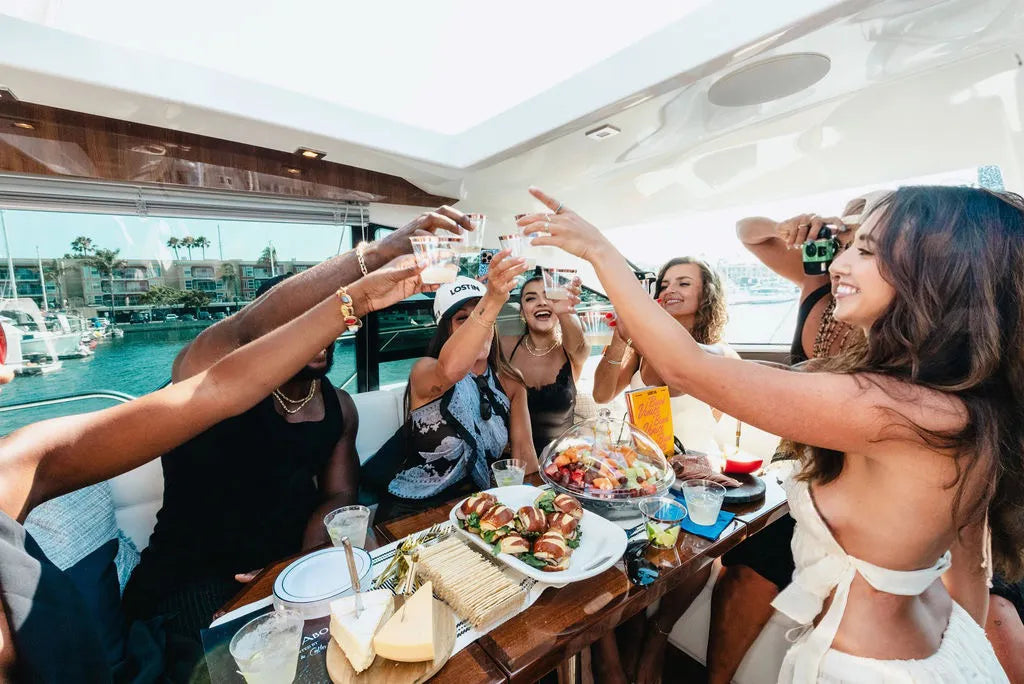 Group of friends toasting drinks on a yacht with food and waterfront view.