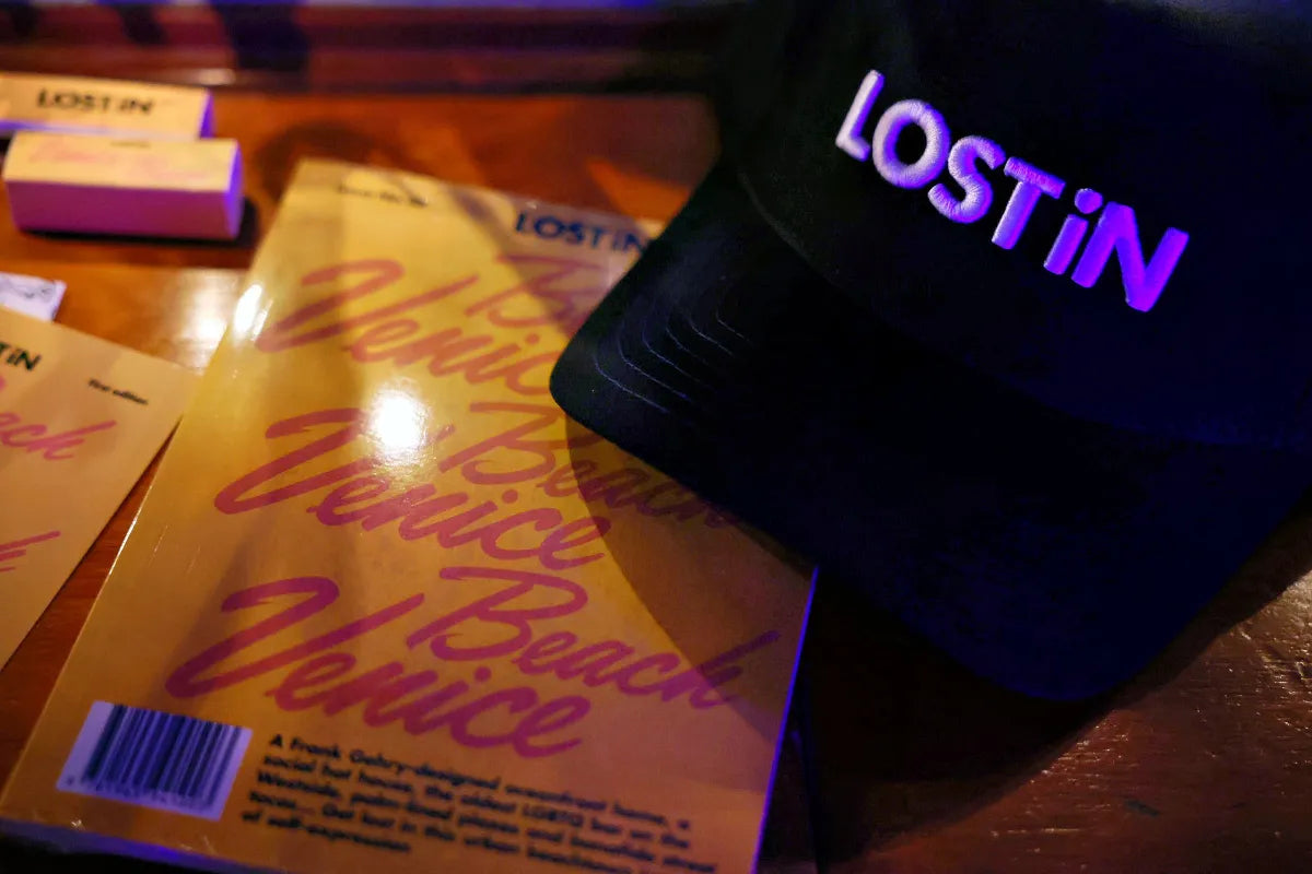 LOST iN Venice Beach guidebook and branded hat on wooden table.